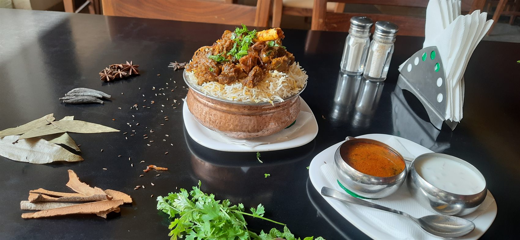 Copper handi of rice topped with mutton curry, rasam and raita alongside