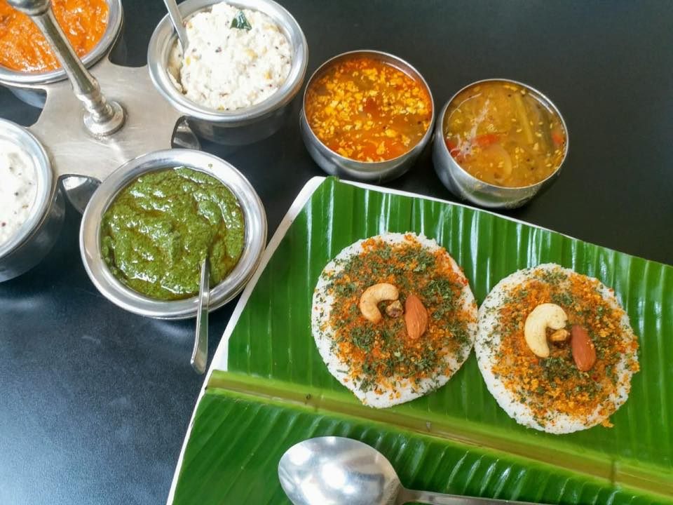 Two idlys topped with podi and cashew on banana leaf, chutney carousel behind